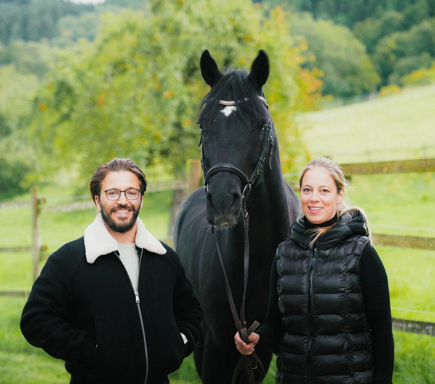 Anna-Lisa Stotmeister zusammen mit ihrem Mann Julian Ehrle in der Mitte ein schwarzes Pferd. Beide lachen in die Kamera.